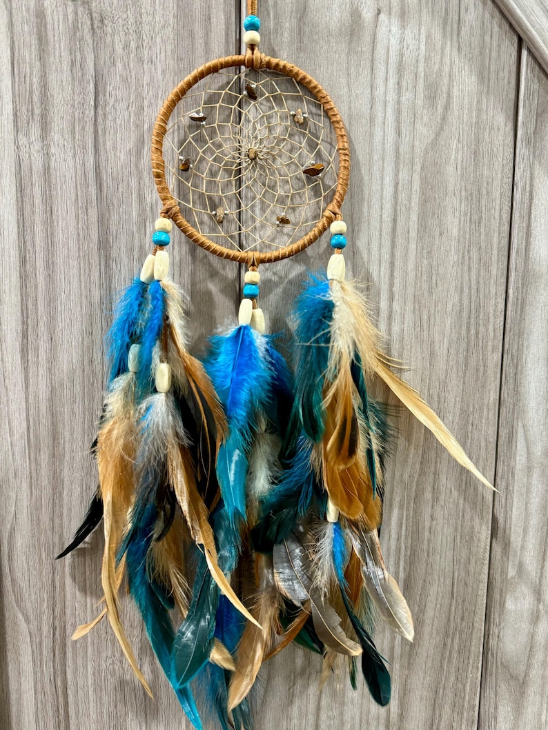 May include: A handcrafted dreamcatcher with a circular, woven web design. The dreamcatcher features a brown wooden hoop, adorned with small stones and hanging feathers in shades of blue, teal, and tan. Decorative beads are also included.