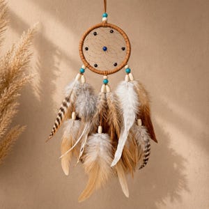 Puede incluir: Un atrapasueños artesanal con un marco circular marrón tejido, adornado con plumas en tonos marrón, blanco y gris. El atrapasueños presenta pequeñas cuentas negras alrededor del marco y cuentas turquesas. Se utiliza un cordón de cuero para colgarlo.