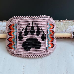 May include: A light pink beaded cuff bracelet with a black bear paw design. The bracelet has orange, turquoise, and black bead accents. A wooden dowel is threaded through the centre, with beaded embellishments on the ends.