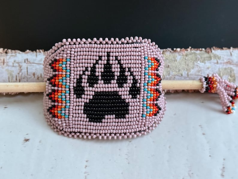 May include: A light pink beaded cuff bracelet with a black bear paw design. The bracelet has orange, turquoise, and black bead accents. A wooden dowel is threaded through the centre, with beaded embellishments on the ends.