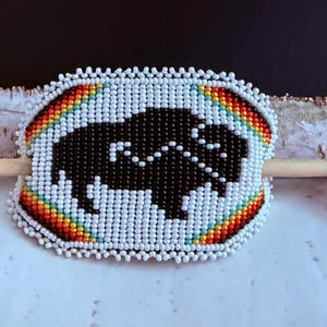 May include: A beaded hair slide with a black buffalo design. The slide has a rainbow-coloured border and white beads. A wooden stick is attached to the back.