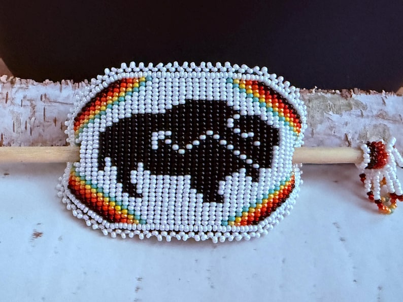 May include: A beaded hair slide with a black buffalo design. The slide has a rainbow-coloured border and white beads. A wooden stick is attached to the back.