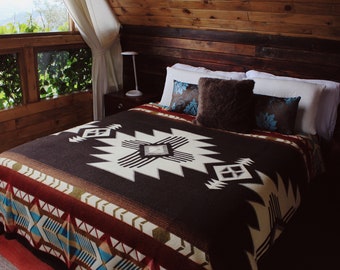 May include: A bed with a brown and white patterned blanket, two decorative pillows, and white pillows. The blanket features a geometric design with red, brown, and cream colours. The room has wooden walls and a window with a view.