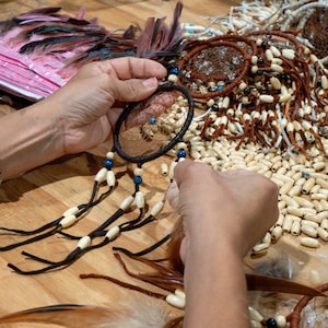 May include: A person is crafting a dreamcatcher. The dreamcatcher has a dark brown circular frame with a woven web and dangling beads. The beads are cream and blue. Feathers and other dreamcatchers are in the background.