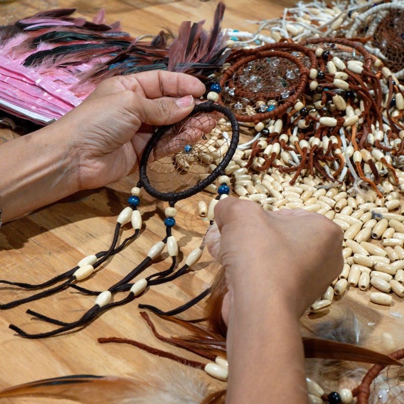 May include: A person is crafting a dreamcatcher. The dreamcatcher has a dark brown circular frame with a woven web and dangling beads. The beads are cream and blue. Feathers and other dreamcatchers are in the background.