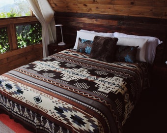 May include: A bed featuring a brown, cream, and blue patterned blanket with geometric shapes and teepee motifs. The bed includes two white pillows, two dark brown pillows with blue floral accents, and a central dark brown faux fur pillow.