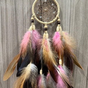 May include: A beige dreamcatcher with a woven web design, adorned with small stones. It features hanging feathers in shades of pink, brown, black, and tan, accented with wooden beads. The dreamcatcher is suspended by a natural-colored cord.