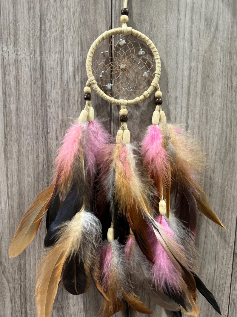 May include: A beige dreamcatcher with a woven web design, adorned with small stones. It features hanging feathers in shades of pink, brown, black, and tan, accented with wooden beads. The dreamcatcher is suspended by a natural-colored cord.