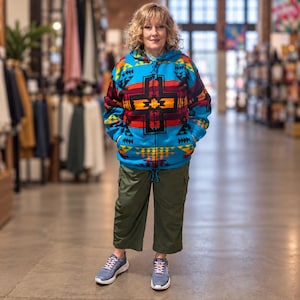 Puede incluir: Una sudadera con capucha y cremallera de color azul brillante con un llamativo patrón geométrico multicolor en rojo, naranja, amarillo y negro. La sudadera incluye capucha con cordón, dos bolsillos delanteros, pantalones cargo cortos verde oliva y zapatillas azules.