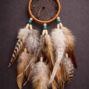 May include: A handcrafted dreamcatcher with a brown leather-wrapped hoop, adorned with feathers in shades of brown, white, and gray. The dreamcatcher features turquoise and wooden beads, and a central web design.