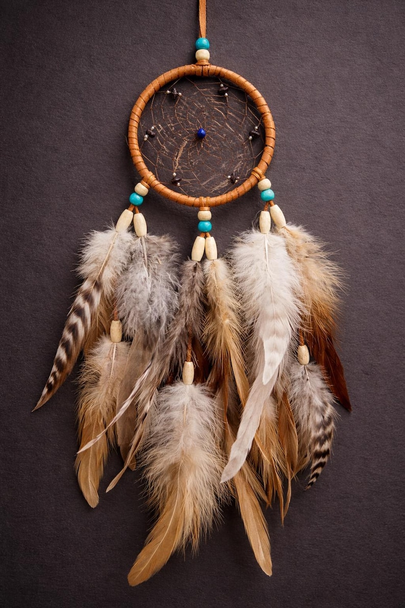 May include: A handcrafted dreamcatcher with a brown leather-wrapped hoop, adorned with feathers in shades of brown, white, and gray. The dreamcatcher features turquoise and wooden beads, and a central web design.