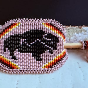 May include: A beaded hair stick with a black buffalo design. The hair accessory is surrounded by pink beads with a colourful border of orange, yellow, and red. The stick is made of wood and has a beaded end.