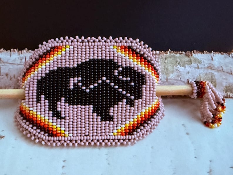 May include: A beaded hair stick with a black buffalo design. The hair accessory is surrounded by pink beads with a colourful border of orange, yellow, and red. The stick is made of wood and has a beaded end.