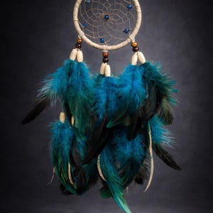May include: A handcrafted dreamcatcher with a beige hoop, intricate web design, and blue beads. The dreamcatcher features vibrant teal and black feathers, and wooden beads. This decorative item is designed to be hung.