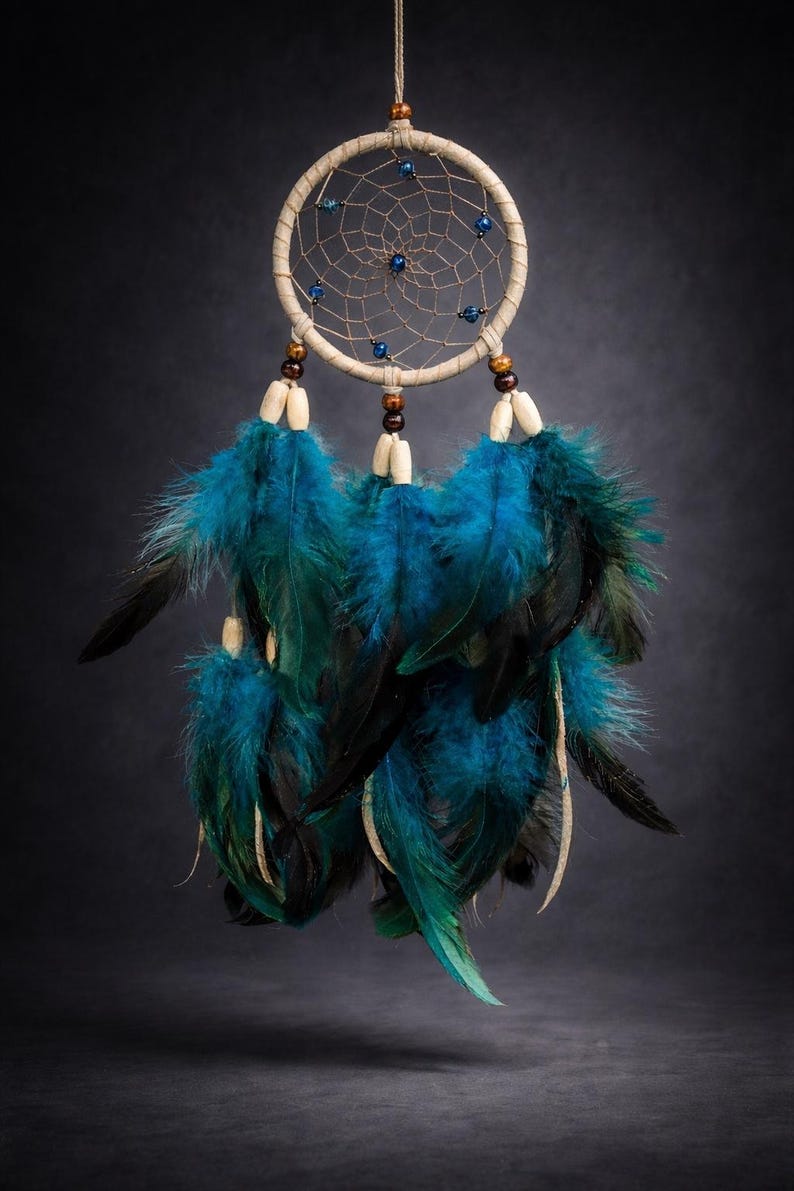 May include: A handcrafted dreamcatcher with a beige hoop, intricate web design, and blue beads. The dreamcatcher features vibrant teal and black feathers, and wooden beads. This decorative item is designed to be hung.