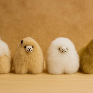 May include: Four fluffy alpaca figurines in shades of cream, tan, and gold are arranged in a row. Each alpaca has a small, black face and a smiling expression. The figurines are on a light brown surface.