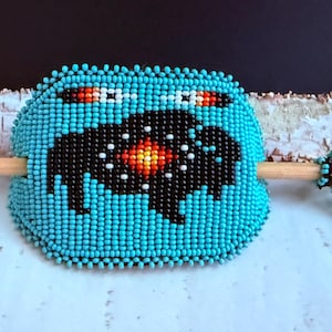 May include: A turquoise beaded hair barrette featuring a buffalo design. The buffalo is black with a red, orange, and white centre. The barrette is on a wooden stick, with a small beaded figure attached.