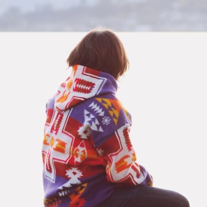 May include: A person wearing a colorful patterned hooded sweater sits on a wooden post, looking out at a body of water.
