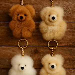 Puede incluir: Cuatro llaveros de osos de peluche esponjosos en tonos marrón, crema y blanco, cada uno con un anillo y una cadena dorados. Los osos tienen ojos y narices de botón negros. El fondo es una superficie de madera.