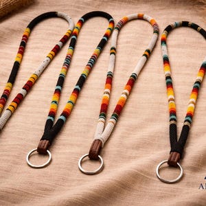 May include: Four beaded lanyards with colorful patterns in black, white, red, yellow, and blue. Each lanyard has a leather accent and a silver ring. The lanyards are displayed on a tan fabric surface. The logo for Amerinkas is in the bottom right corner.