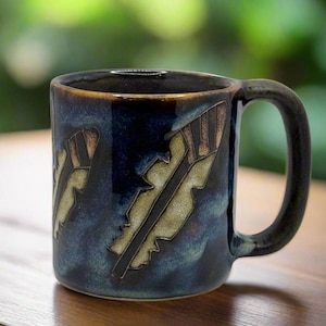 Pode incluir: Uma caneca de cerâmica azul com uma alça. A caneca tem um design de penas estilizadas em tons de marrom e branco.