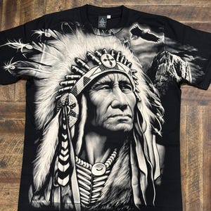May include: Black t-shirt with a white and gray graphic print of a Native American chief wearing a headdress. The chief is looking to the right and has a serious expression on his face. The background of the graphic includes a mountain and a flying eagle.