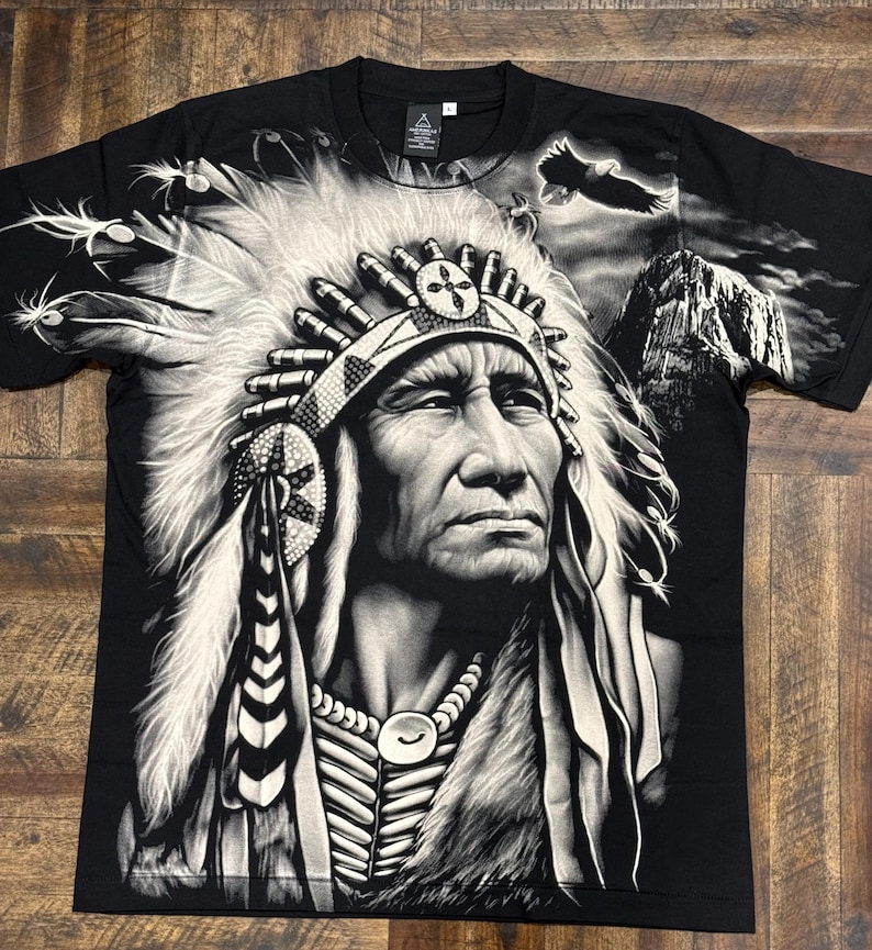 May include: Black t-shirt with a white and gray graphic print of a Native American chief wearing a headdress. The chief is looking to the right and has a serious expression on his face. The background of the graphic includes a mountain and a flying eagle.