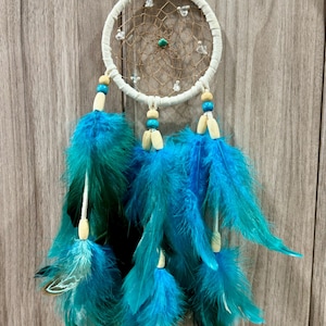 May include: A handcrafted dreamcatcher with a white circular frame and a woven web. It features turquoise and blue feathers, wooden beads, and small clear beads. The dreamcatcher is designed for wall decor.