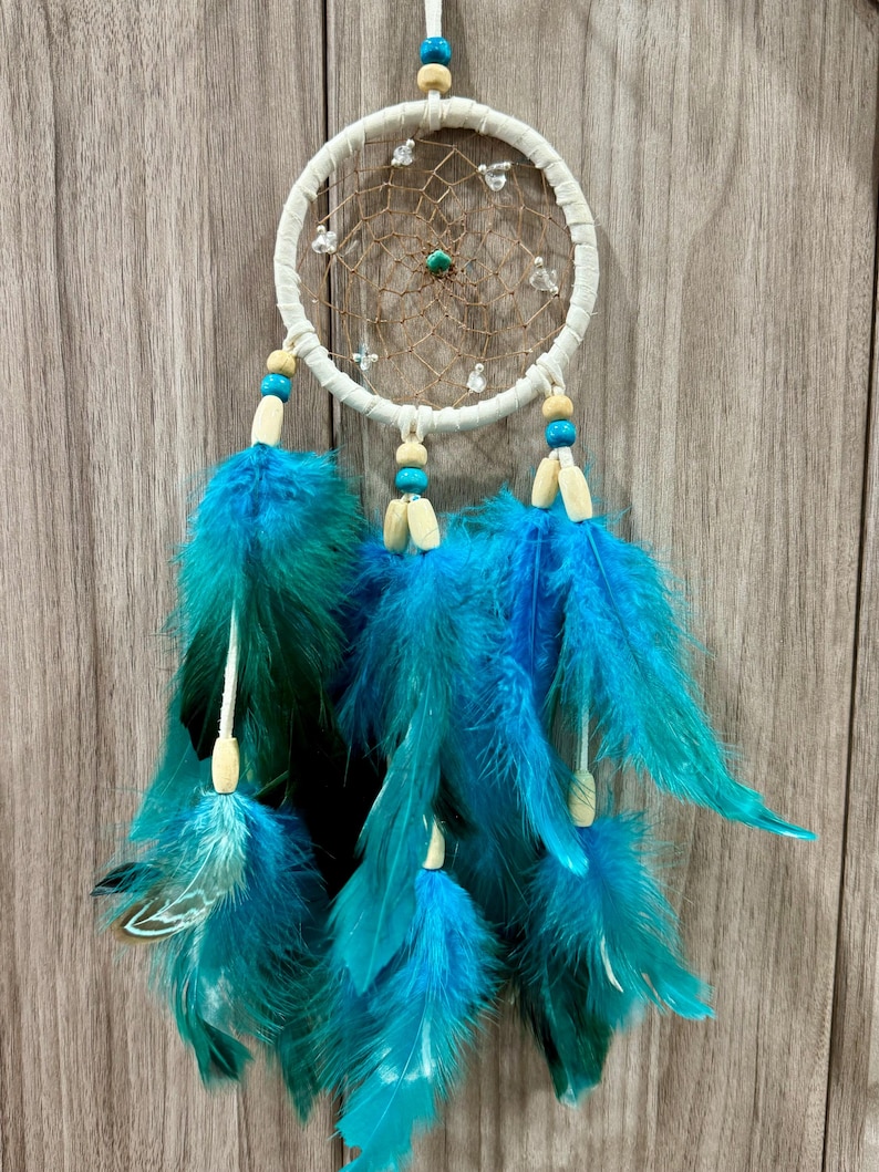 May include: A handcrafted dreamcatcher with a white circular frame and a woven web. It features turquoise and blue feathers, wooden beads, and small clear beads. The dreamcatcher is designed for wall decor.