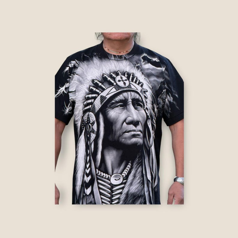 May include: Black and white graphic t-shirt featuring a Native American chief wearing a headdress with feathers. The chief's face is detailed and the background is a blurred image of a cloudy sky.