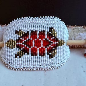 May include: A beaded hair barrette with a turtle design. The barrette is white with a red and black turtle design in the centre. The barrette is approximately 7.6 cm wide. A small beaded accent piece is next to the barrette.