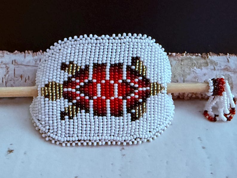 May include: A beaded hair barrette with a turtle design. The barrette is white with a red and black turtle design in the centre. The barrette is approximately 7.6 cm wide. A small beaded accent piece is next to the barrette.