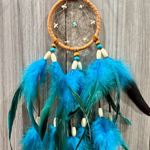 May include: A handcrafted dreamcatcher with a tan-colored circular frame and a woven web design. It features turquoise and wooden beads, small white accents, and vibrant blue feathers hanging below. The dreamcatcher is suspended by a leather cord.