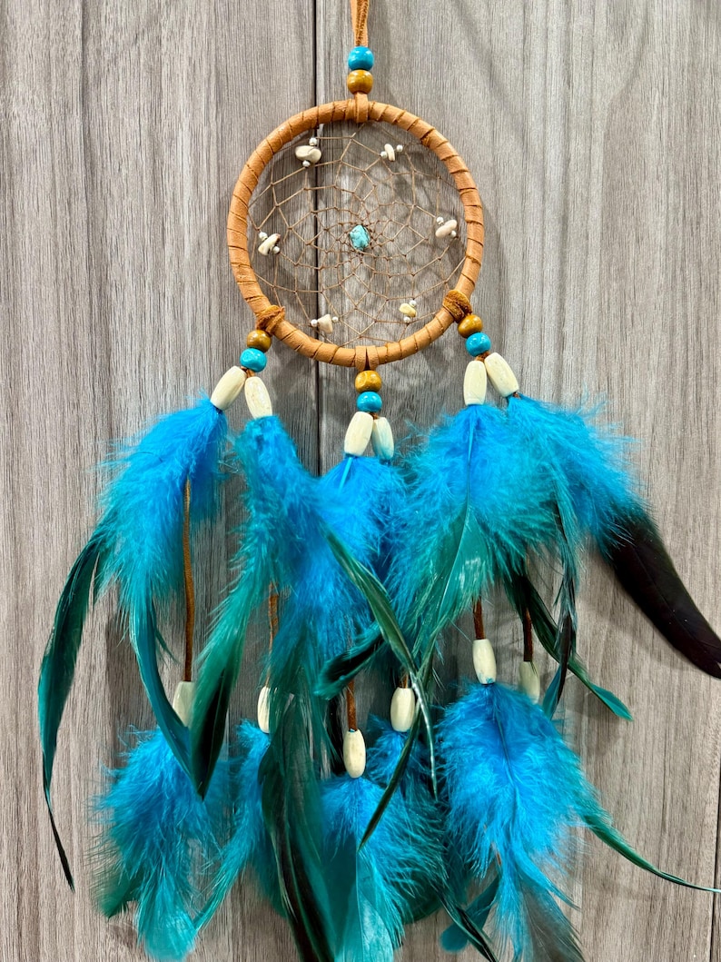 May include: A handcrafted dreamcatcher with a tan-colored circular frame and a woven web design. It features turquoise and wooden beads, small white accents, and vibrant blue feathers hanging below. The dreamcatcher is suspended by a leather cord.