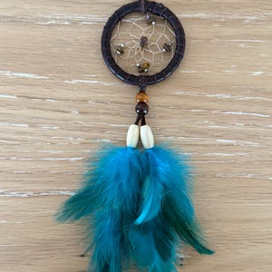 Puede incluir: Un atrapasueños azul turquesa con cuero marrón y cuentas de madera. El atrapasueños tiene un marco circular con una red de cuerda y piedras marrones.