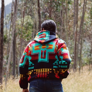 May include: A person wearing a colourful hooded sweater with a geometric pattern in shades of red, blue, yellow, and black. The sweater has a hood and is worn over a pair of blue jeans.