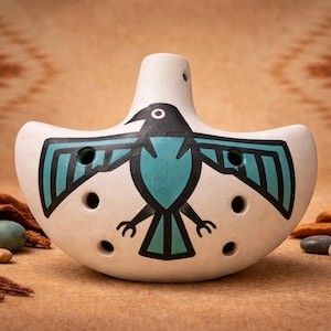 Può includere: Ocarina in ceramica color crema con un disegno di uccello turchese e nero dipinto. L'uccello ha le ali spiegate ed è centrato sull'ocarina. Lo strumento ha più fori per le dita e un bocchino. Lo sfondo è un tessuto beige a fantasia.