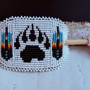 May include: A white beaded hair barrette featuring a black bear paw print and colourful blue, yellow, and red accents. A smaller beaded piece is attached to a wooden stick. The barrette is a hair accessory.