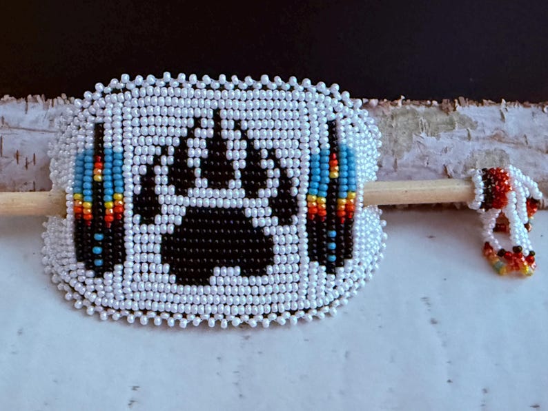 May include: A white beaded hair barrette featuring a black bear paw print and colourful blue, yellow, and red accents. A smaller beaded piece is attached to a wooden stick. The barrette is a hair accessory.