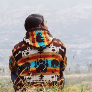 Pode incluir: Uma pessoa vestindo um suéter estampado em marrom, laranja, azul e branco com um design de pata de urso. A pessoa está sentada em um campo de grama alta e olhando para a distância.