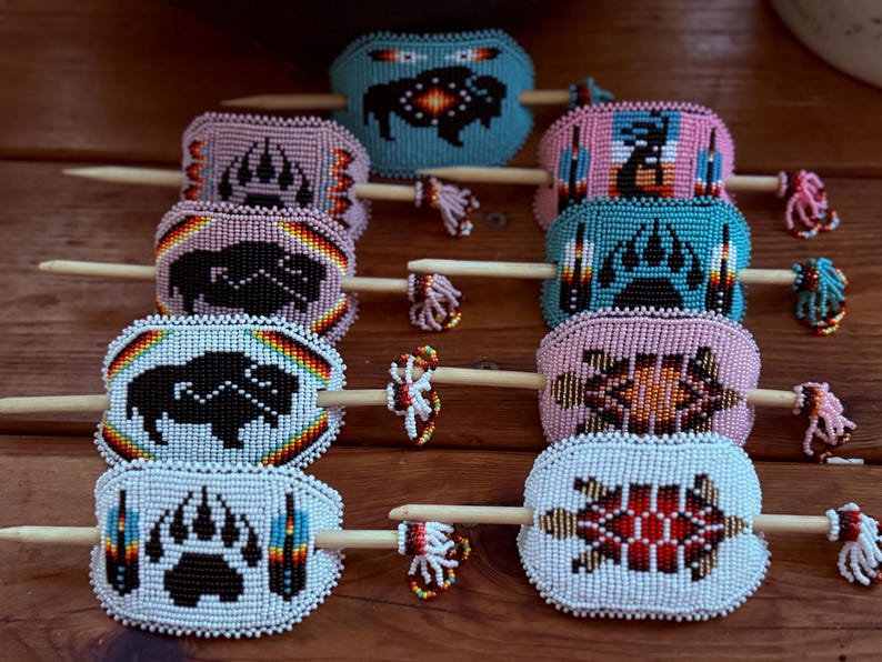 May include: A selection of beaded hair sticks with diverse designs. The hair sticks showcase colourful beadwork, featuring images of a bear paw, buffalo, and geometric patterns. The sticks are made of wood and are displayed on a wooden surface.