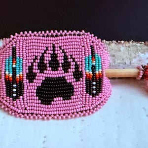 May include: Pink beaded hair accessory with a black bear paw print. It features a wooden stick for insertion and is decorated with beaded feather designs in turquoise, red, and white. The stick's end is adorned with pink beads.
