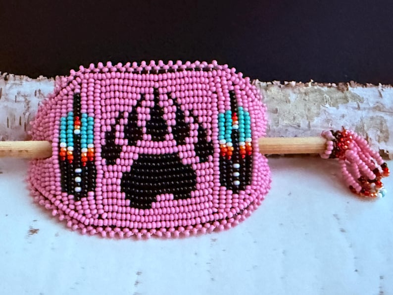 May include: Pink beaded hair accessory with a black bear paw print. It features a wooden stick for insertion and is decorated with beaded feather designs in turquoise, red, and white. The stick's end is adorned with pink beads.