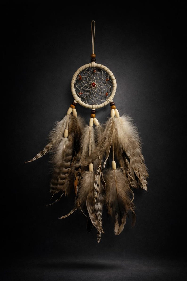 May include: A beige dreamcatcher with a woven web design and brown feathers. The dreamcatcher has a circular frame with a central red accent and wooden beads. The feathers are various shades of brown and beige.