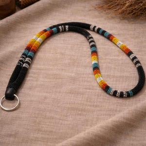 May include: A lanyard crafted from small, round beads in a tubular shape. The design features a repeating pattern of black, white, yellow, orange, red, and turquoise beads. A silver-toned metal ring is attached to one end of the beaded cord.