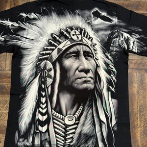 May include: Black t-shirt with a graphic print of a Native American chief wearing a headdress. The chief is looking to the right and has a serious expression on his face. The background is a dark, cloudy sky with a mountain in the distance.