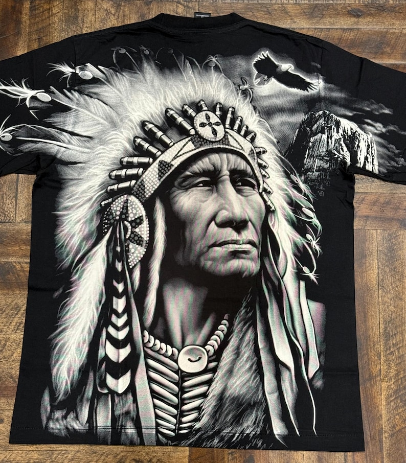 May include: Black t-shirt with a graphic print of a Native American chief wearing a headdress. The chief is looking to the right and has a serious expression on his face. The background is a dark, cloudy sky with a mountain in the distance.