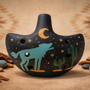 Può includere: Ocarina in ceramica nera con un design a tema deserto. Lo strumento presenta un lupo che ulula, cactus, una luna crescente e stelle. L'ocarina ha diversi fori per le dita e un bocchino.