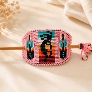 May include: Pink beaded hair barrette with a Southwestern design. The barrette features a central figure with black, orange, and white details, flanked by turquoise and black shapes. A wooden stick secures the barrette. A small beaded accent hangs from the side.