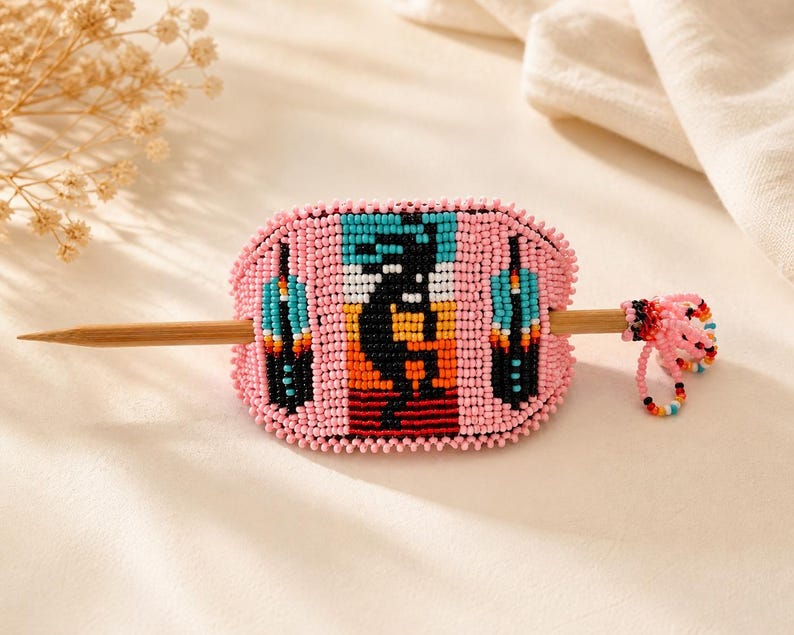 May include: Pink beaded hair barrette with a Southwestern design. The barrette features a central figure with black, orange, and white details, flanked by turquoise and black shapes. A wooden stick secures the barrette. A small beaded accent hangs from the side.